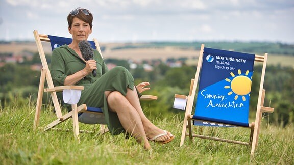 Die Moderatorin Sandra Voigtmann sitzt auf einem Liegestuhl mit der Aufschrift „Sonnige Aussichten“ in einer Wiese