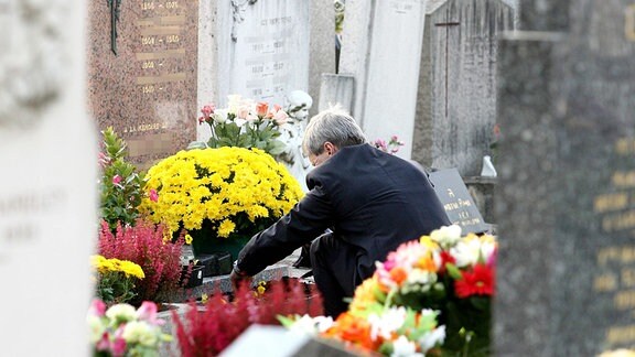 Auf einem Friedhof, ein Mann hockt vor einem Grab und bringt Blumen.