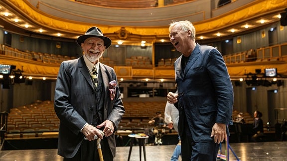Zwei Männer lachen auf einer Opernbühne. Im Hintergrund: Blick in den leeren Zuschauerraum.