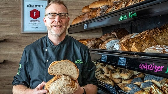 Bäckermeister Silberbach in seinem Verkaufsraum, mit aufgeschnittenem Brot in der Hand, freudig lächelnd