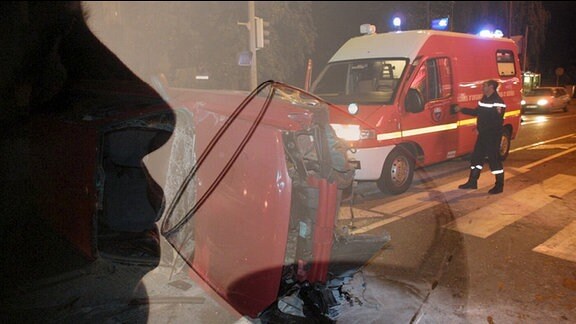 Im Vordergrund eine Frau, die aus einem Weinglas trinkt und im Hintergrund ein Autounfall mit Rettungswagen.