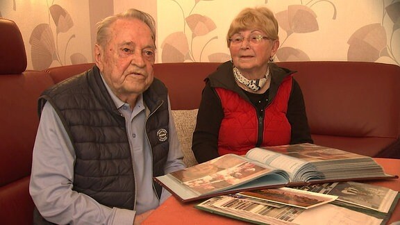 Ein älteres Paar sitzt an einem Tisch, auf dem ein Fotoalbum liegt.