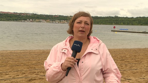 Eine Frau steht vor einem See und spricht in die Kamera.