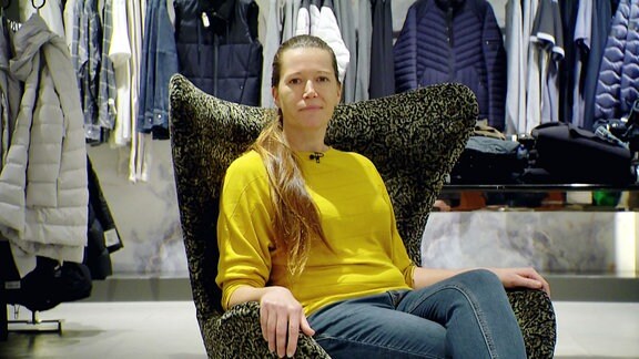 Eine Frau in schlichter Kleidung, gelber Pulli und Jeans, sitzt in einem Sessel und schaut in die Kamera.
