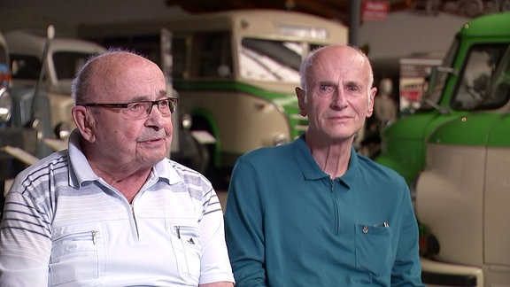 Zwei ältere Männer sitzen während eines Interviews in einer großen Halle, im Hintergrund verschiedene Fahrzeuge des Typs Barkas.