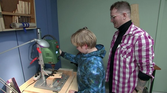 Ein Junge probiert sich an einer stationären Bohrmaschine, ein Mann hilft ihm dabei.