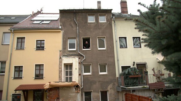 Ein schmales graues Haus zwischen breiteren Häusern, ein Mann hat ein Fenster geöffnet.