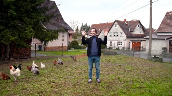 Reporter Stefan Ganß steht auf einer Wiese neben Hühnern und winkt mit den Händen.