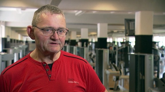 Ein älterer Mann mit Brille, kurzen Haaren und rotem T-Shirt spricht während eines Interviews. Im Hintergrund Sportgeräte im Fitnessstudio.