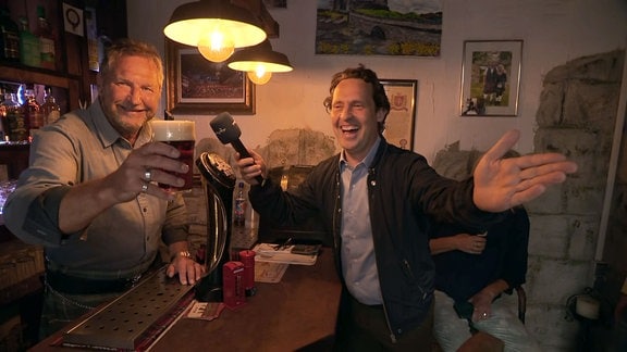 Reporter Stefan Ganß lacht mit einem Mann der ein Bierglas in der Hand hält.