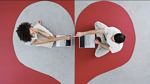 Symbolbild: Auf einem Teppich, der ein Herz zeigt, sitzen sich eine Frau und ein Mann mit Laptop gegenüber, deren Zeigefinger sich berühren