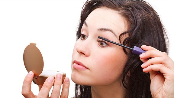Eine Junge Frau trägt Wimperntusche auf und sieht in einen Taschenspiegel