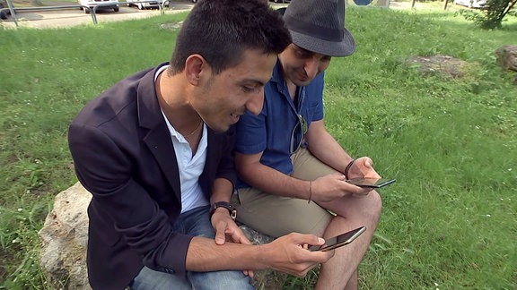 Zwei Männer sitzen auf einem Stein auf einer Grünfläche und halten ihre Smartphones in der Hand