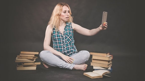 Eine junge Frau sitzt umgeben von Büchern auf dem Boden und filmt sich mit einem Smartphone.