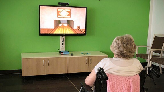 Eine Seniorin sitzt in einem Rollstuhl vor einem Bildschirm und spielt ein Videospiel.