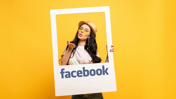 Eine Frau blickt durch einen weißen Rahmen, auf dem "facebook" steht und wirft der Kamera einen Kuss zu.