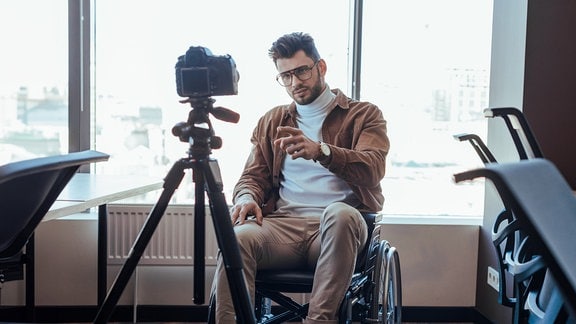 Ein Mann im Rollstuhl spricht in eine Kamera auf einem Stativ.