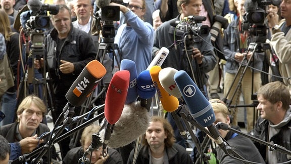 Kamerateams und Fotografen warten vor einer Pressekonferenz hinter Mikrofonen.
