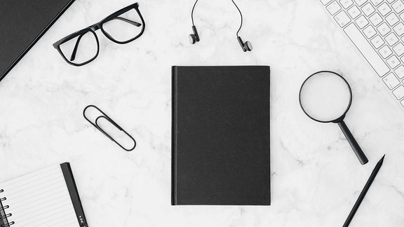 Ein schwarzes Tagebuch mit Brille, Kopfhörer, Lupenbrille, Bleistift und Tastatur auf einem Schreibtisch