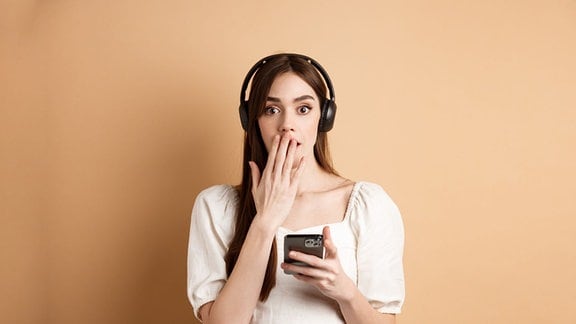 Eine Frau mit Köpfhörern auf dem Kopf und einem Smartphone in der Hand hält sich erschrocken die Hand vor den Mund.