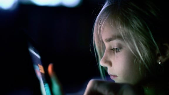 Ein junges Mädchen blickt auf einen Handybildschirm. Ihr Gesicht wird vom Bildschirm angeleuchtet.