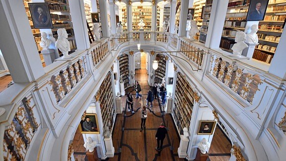 Besucher besichtigen den Rokokosaal der Herzogin Anna Amalia Bibliothek. Weitwinkel-Ansicht. Mittig am Geländer, viele Säule, weiß und gold dominieren, viele Bücher hinter den Säulen.