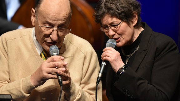 Manfred Krug und Uschi Brüning stehen auf einer Bühne und singen in ihr Mikrofon.