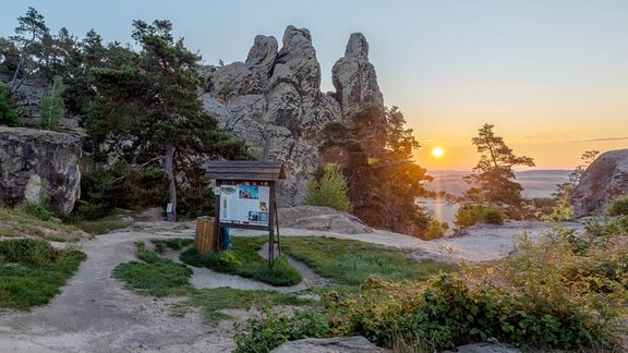 Die Teufelsmauer, eine Sandstein-Formation, mehrere Wege und eine Informationstafel im Vordergrund, im Hintergrund die untergehende Sonne. 