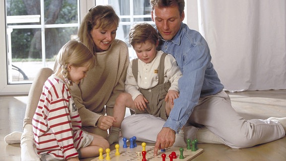 Familie mit zwei Kindern sitzt auf dem Fußboden vor dem Fenster und spielt eine Partie Mensch-ärgere-dich-nicht