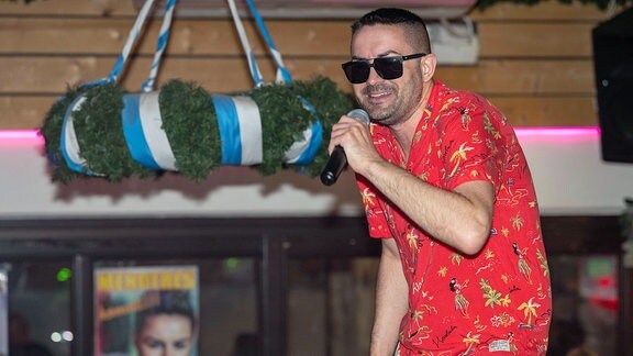 Ein Mann mit Sonnenbrille und rotem Hawaiihemd singt auf einer Bühne.