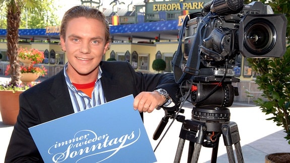 Der Volksmusiker Stefan Mross lächelt neben einer Fernsehkamera und mit dem Logo der ARD-Unterhaltungssendung "Immer wieder sonntags" in die Kamera.