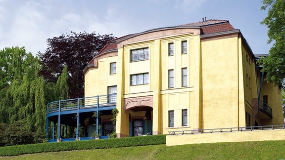  Blick von der Straße auf eine zweigeschossige moderne Villa mit gelber Fassade auf einem begrünten Hügel. 