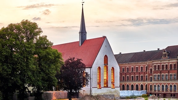 Klosterkirche Grimma, ein Gebäude mit roten Dach, einem Turm und drei großen Fenstern, auf der Linken seite ein Baum, rechts im Hintergrund Häuser. 