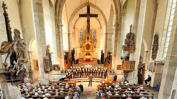 Innenansicht des Merseburger Doms: Kirchenschiff mit gewölbter Decke, rechts und link Sitzbänke, über dem Altar ein Kreuz