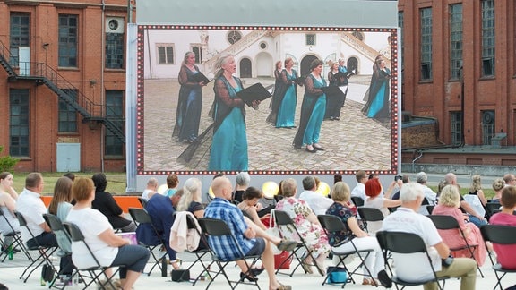 Eröffnungskonzert MDR-Musiksommer in Hoyerswerda: Auf einer Videoleinwand hinter dem Publikum ist der MDR-Rundfunkchor zu sehen.