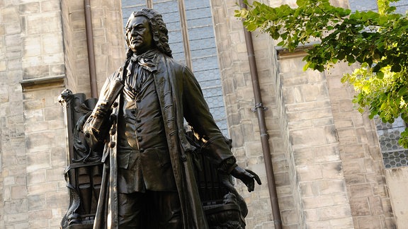 Eine Statue eines Mannes mit langen, lockigen Haaren und historischem Gewand steht vor einer Kirche.
