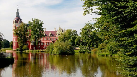 Blick über einen See auf ein Schloss mit roter Fassade hinter grünen Bäumen.