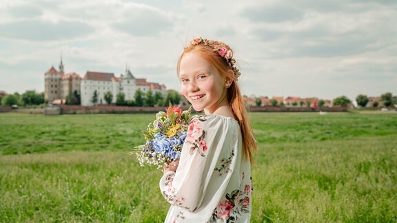 Motiv der Sächsischen Landesgartenschau Torgau 2022: Ein Mädchen steht mit einem Blumenstrauß auf den Elbauen in Torgau, im Hintergrund Schloss Hartenfels.