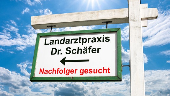 Ein weisser Holzgalgen mit einem grün gerahmten weißen Schild mit der Aufschrift "Landarztpraxis Dr. Schaefer Nachfolger gesucht" sowie einem Richtungspfeil vor einem blauen Himmel mit Schleierwolken und Sonne.