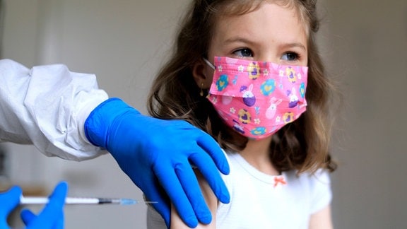 Ein Mädchen mit einer rosafarbenen Maske bekommt von einem Arzt eine Spritze