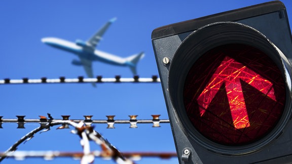 Stacheldraht und rotes Ampellicht vor startendem Flugzeug.