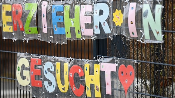 Mit bunten Buchstaben sind die Worte „Erzieher*in gesucht“ geformt, die an einem Zaun einer Kindertagesstätte im Stadtteil Sachsenhausen befestigt sind.