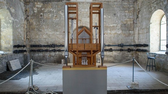 Ein Holzgestell mit wenigen Orgelpfeifen in einem leeren Kirchenraum, bei dem die Wände ohne Putz sind.
