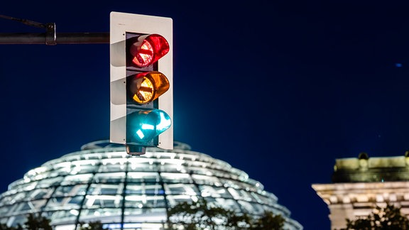 Eine Ampel vor dem Reichstagsgebäude leuchtet in einer Langzeitbelichtung in allen drei Phasen, wobei sich der Straßenverkehr als Leuchtspuren abzeichnet.