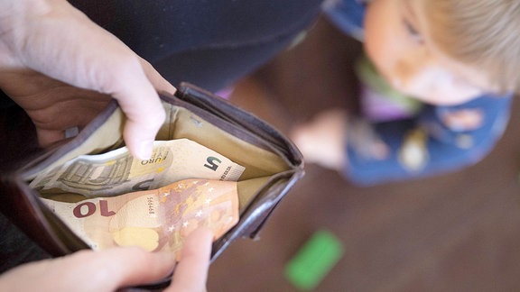 Ein Kind schaut hoch, wo eine Hand ein Geldschein aus einem Portemonnaies nimmt.