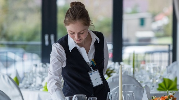 Eine Restaurantfachfrau kontrolliert an einem eingedeckten Tisch die Gläser