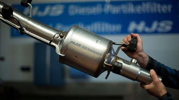 Ein Mitarbeiter hält einen SCR-Katalysator zur Nachrüstung für Diesel zur Stickstoffreduzierung bei Firma HJS in der Werkstatt hoch