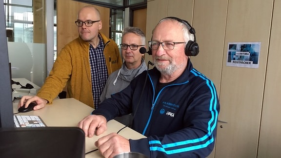 Drei Männer stehen an einem Pult und schauen auf einen Computerbildschirm. Einer der Männer trägt Kopfhörer, ein anderer bedient die Maus