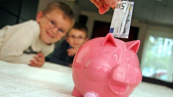 Zwei Jungs blicken auf ein pinkes Sparschwein, welches mit einem 5-Euro-Schein gefüttert wird