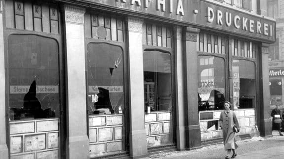 November 1938, eine Frau läuft an einer Druckerei mit kaputten Fensterscheiben vorbei.